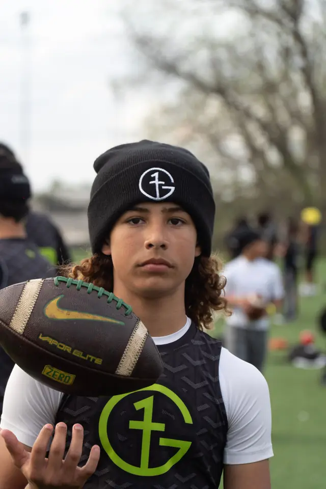 A young G1 Football Academy athlete holding a football, wearing a G1 crest pinnie and team beanie