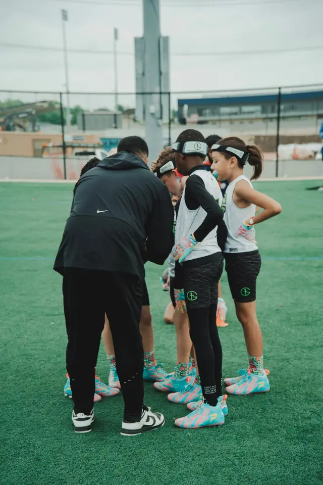 A G1 coach crouched low inside a huddle of young athletes in white and teal kit